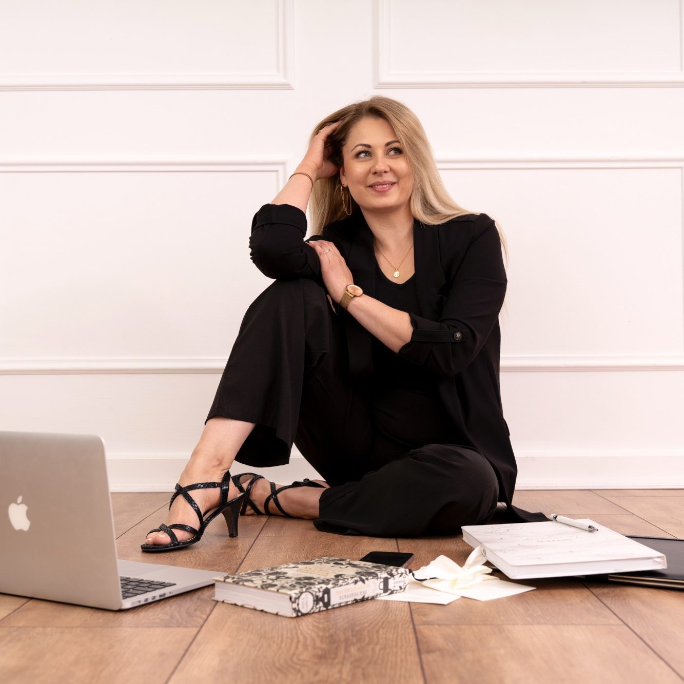 Frau sitzt lächelnd mit Laptop und Notizen auf dem Boden, in eleganter Kleidung.