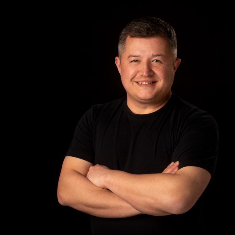 Männlicher Mann mit gekreuzten Armen, lächelnd, in schwarzem Shirt vor schwarzem Hintergrund.